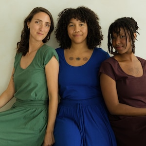 May include: Three women wearing solid-colored dresses. The dresses are in shades of green, blue, and burgundy. The dresses have a scoop neckline and short sleeves. The women are sitting in front of a white wall.