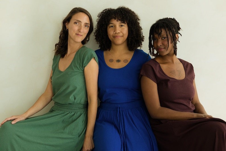 May include: Three women wearing solid-colored dresses. The dresses are in shades of green, blue, and burgundy. The dresses have a scoop neckline and short sleeves. The women are sitting in front of a white wall.