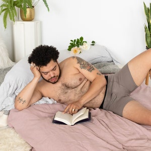 Puede incluir: Una persona tumbada en una cama leyendo un libro. Lleva pantalones cortos de boxeo grises y tiene un tatuaje en el brazo. La cama tiene una s&aacute;bana de lino rosa y una almohada blanca y esponjosa.
