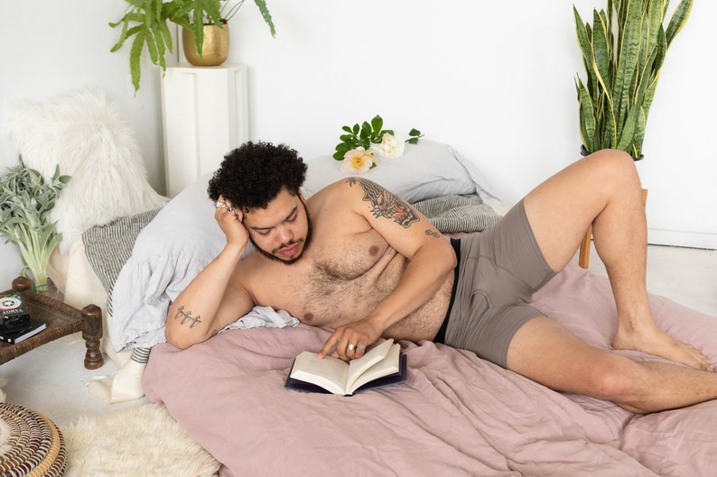 Puede incluir: Una persona tumbada en una cama leyendo un libro. Lleva pantalones cortos de boxeo grises y tiene un tatuaje en el brazo. La cama tiene una s&aacute;bana de lino rosa y una almohada blanca y esponjosa.