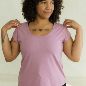 May include: A mauve scoop-neck t-shirt with short sleeves. The shirt is made of a soft, heathered fabric. The model is wearing black pants. The background is a plain, off-white wall.