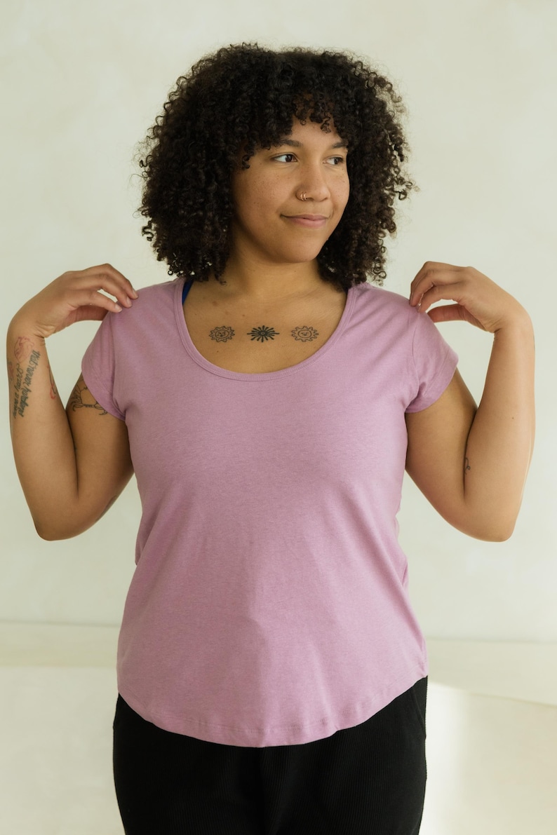 May include: A mauve scoop-neck t-shirt with short sleeves. The shirt is made of a soft, heathered fabric. The model is wearing black pants. The background is a plain, off-white wall.