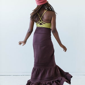 May include: A woman wearing a maroon maxi skirt with a ruffled hem, a green tank top with a criss-cross back, and a pink beanie.