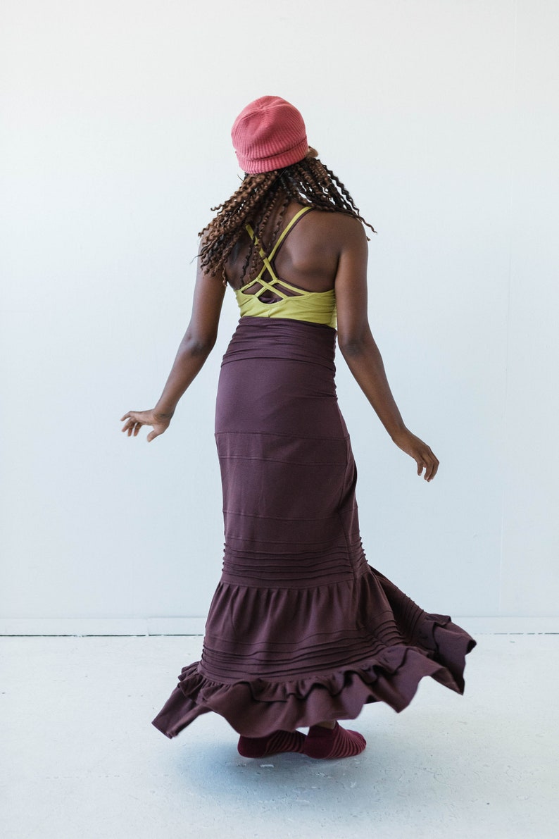 May include: A woman wearing a maroon maxi skirt with a ruffled hem, a green tank top with a criss-cross back, and a pink beanie.