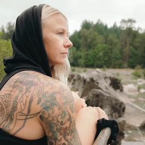 May include: A woman with blonde hair wearing a black hooded top and black pants. She has a large tattoo on her left arm and a smaller tattoo on her right arm. She is sitting on a rock with a river in the background.
