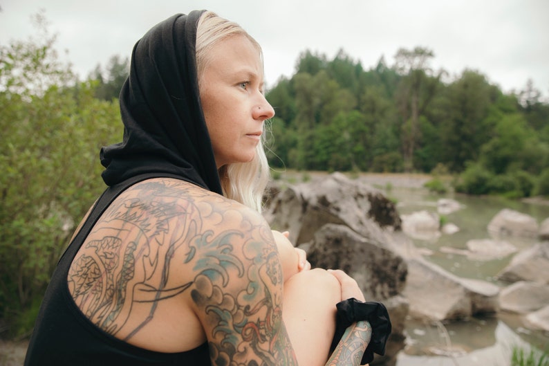 May include: A woman with blonde hair wearing a black hooded top and black pants. She has a large tattoo on her left arm and a smaller tattoo on her right arm. She is sitting on a rock with a river in the background.