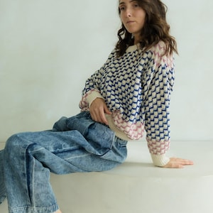 May include: A sweater with a geometric pattern in navy blue, cream, and pink. The sweater is paired with light blue acid wash jeans. The model is sitting on a white surface.