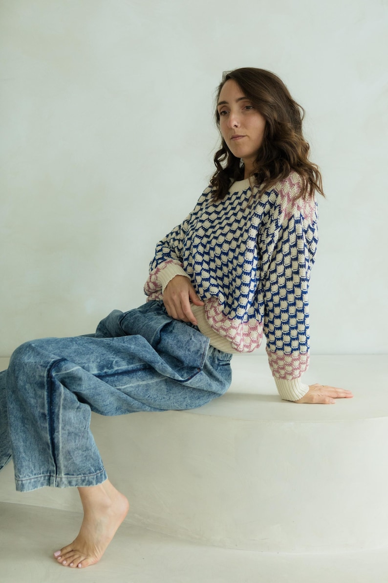 May include: A sweater with a geometric pattern in navy blue, cream, and pink. The sweater is paired with light blue acid wash jeans. The model is sitting on a white surface.