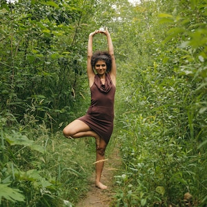 May include: A woman in a brown dress stands in a forest, practicing yoga in a tree pose.