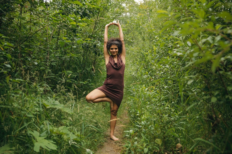 May include: A woman in a brown dress stands in a forest, practicing yoga in a tree pose.