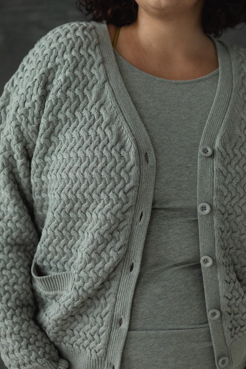 May include: A gray knitted cardigan with a textured pattern and button closure. The cardigan has a relaxed fit and a slightly cropped length.