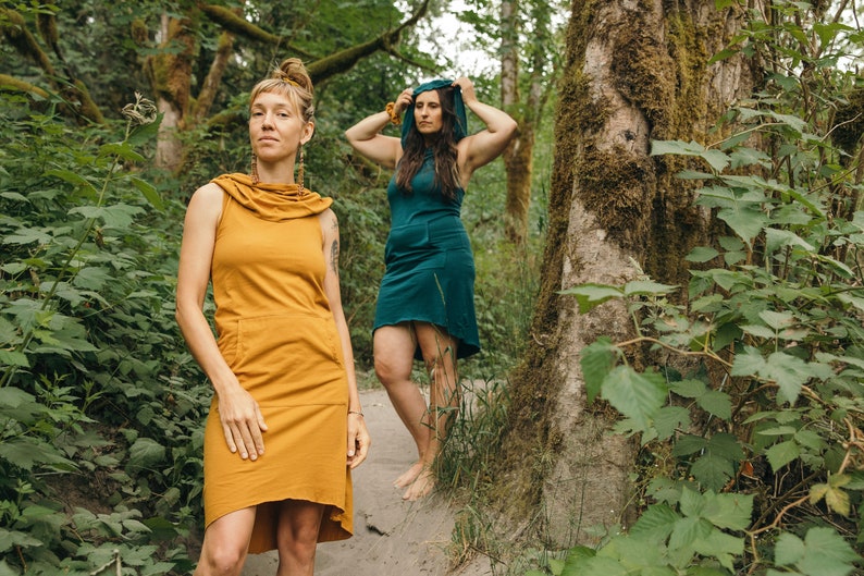 May include: Two people wearing hooded dresses in a forest setting. One dress is mustard yellow and the other is teal.