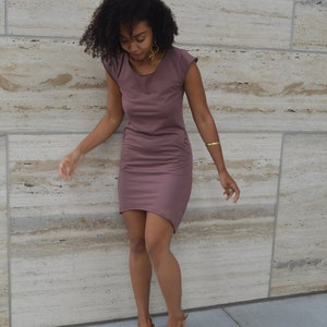 May include: A mauve-colored short-sleeved dress with a round neckline and a high-low hemline. The dress features a simple, solid-colored design with a fitted silhouette that reaches above the knees in the front and slightly longer in the back.