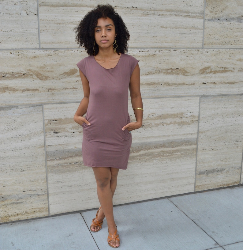 May include: A mauve-colored, short-sleeved dress featuring a round neckline and two front pockets. The garment is made of a solid-colored, lightweight fabric that falls above the knee, providing a simple and functional design for everyday wear.