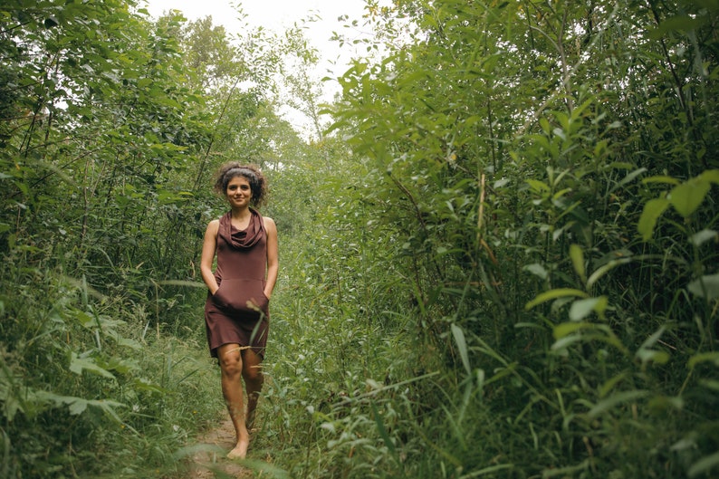 May include: A woman in a brown cowl neck dress walks on a path through a lush green forest.
