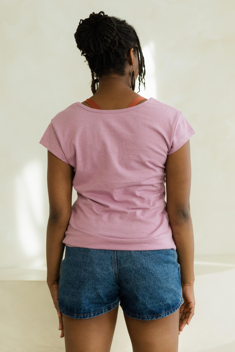 May include: A person wearing a short-sleeved, dusty rose-colored t-shirt and denim shorts. The t-shirt has a scoop neck. The shorts are a medium wash denim. The person has dark hair and is facing away from the camera.
