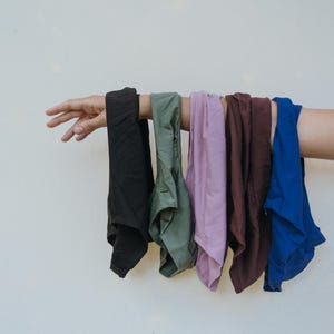 May include: A hand holds up a row of folded garments in various colors. The colors include black, olive green, lavender, maroon, and royal blue. The garments appear to be made of a soft fabric.