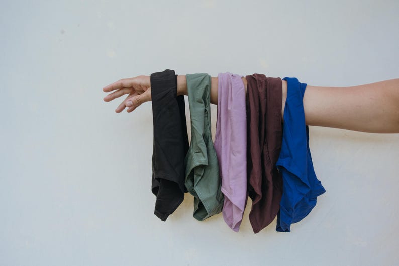 May include: A hand holds up a row of folded garments in various colors. The colors include black, olive green, lavender, maroon, and royal blue. The garments appear to be made of a soft fabric.