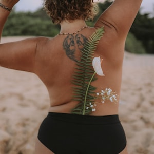 May include: A person's back is adorned with a fern, white flower, and small white flowers. The person is wearing black bottoms. A tattoo is visible on the back. The person is holding leaves above their head.