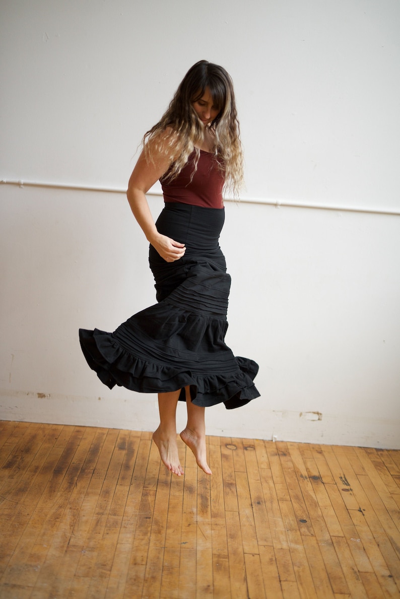 May include: A woman wearing a maroon tank top and a black skirt with multiple ruffles is jumping in the air. The skirt is flowing around her as she jumps. The background is a white wall.