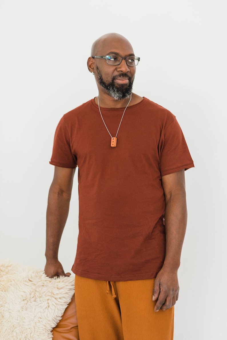 Pu&ograve; includere: Un uomo che indossa una maglietta marrone con una collana a catena d'argento e un piccolo ciondolo rettangolare. Indossa pantaloni arancioni e ha la testa rasata e gli occhiali.