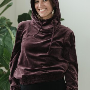 May include: A burgundy velvet hoodie with a drawstring hood and a gathered waist. The long-sleeved top is paired with black trousers. The model is smiling in front of a green plant.