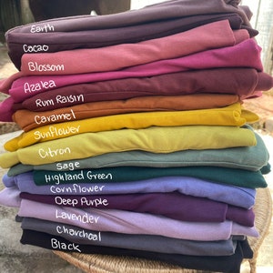 May include: A stack of folded fabric garments in various colors labeled Earth, Cacao, Blossom, Azalea, Rum Raisin, Caramel, Sunflower, Citron, Sage, Highland Green, Cornflower, Deep Purple, Lavender, Charcoal, and Black, arranged in a vertical pile.