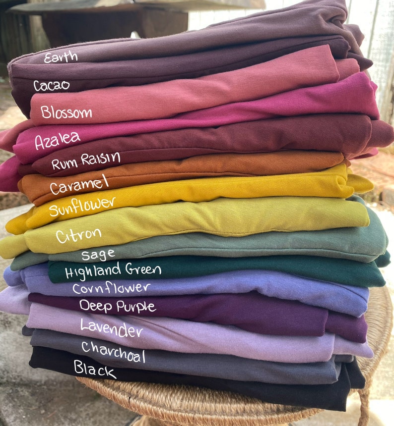 May include: A stack of folded fabric garments in various colors labeled Earth, Cacao, Blossom, Azalea, Rum Raisin, Caramel, Sunflower, Citron, Sage, Highland Green, Cornflower, Deep Purple, Lavender, Charcoal, and Black, arranged in a vertical pile.