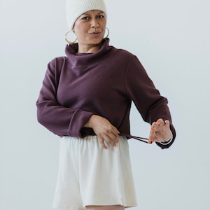 May include: A woman wearing a white beanie, a purple turtleneck sweater, and white shorts. The sweater has a drawstring at the bottom and she is holding the drawstring in her right hand.