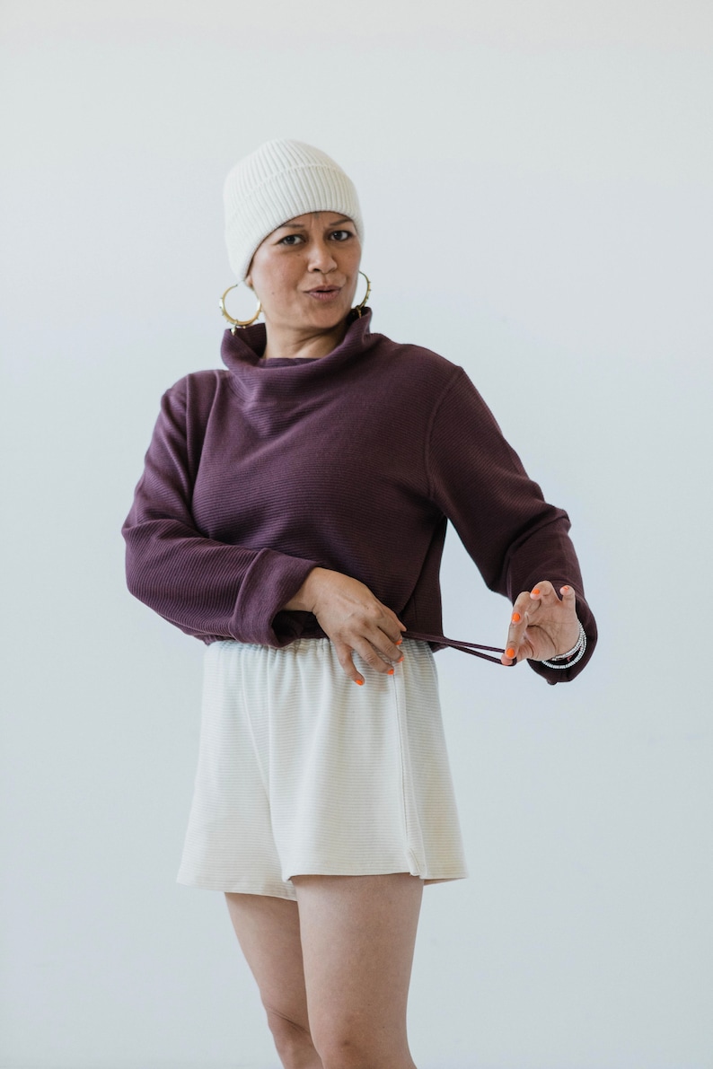 May include: A woman wearing a white beanie, a purple turtleneck sweater, and white shorts. The sweater has a drawstring at the bottom and she is holding the drawstring in her right hand.