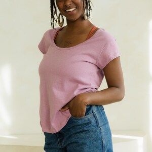 May include: A woman wearing a short-sleeved, scoop-neck mauve t-shirt and denim shorts. She is smiling and has her hand in her pocket. She is wearing gold earrings and has dreadlocks.