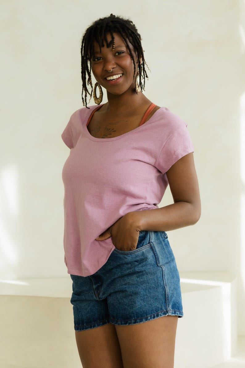 May include: A woman wearing a short-sleeved, scoop-neck mauve t-shirt and denim shorts. She is smiling and has her hand in her pocket. She is wearing gold earrings and has dreadlocks.