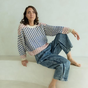 May include: A patterned sweater with a blue and white geometric design, accented with pink and cream details. The sweater is paired with blue denim jeans. The overall aesthetic is casual and comfortable.