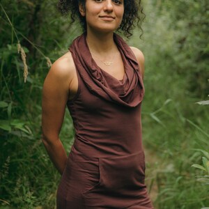May include: A woman wearing a brown cowl neck sleeveless dress with a pocket. She is standing in a forest setting.