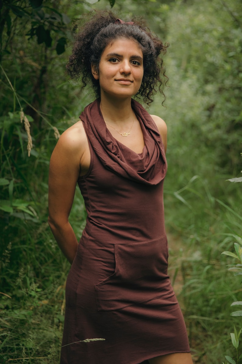 May include: A woman wearing a brown cowl neck sleeveless dress with a pocket. She is standing in a forest setting.