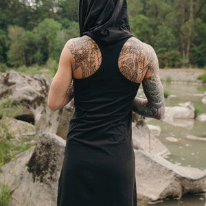 May include: A black hooded tank dress with a scoop back. The dress is made of a soft, comfortable fabric and has a relaxed fit. The model is wearing the dress with a hood pulled up over their head. The model has tattoos on their arms and back.