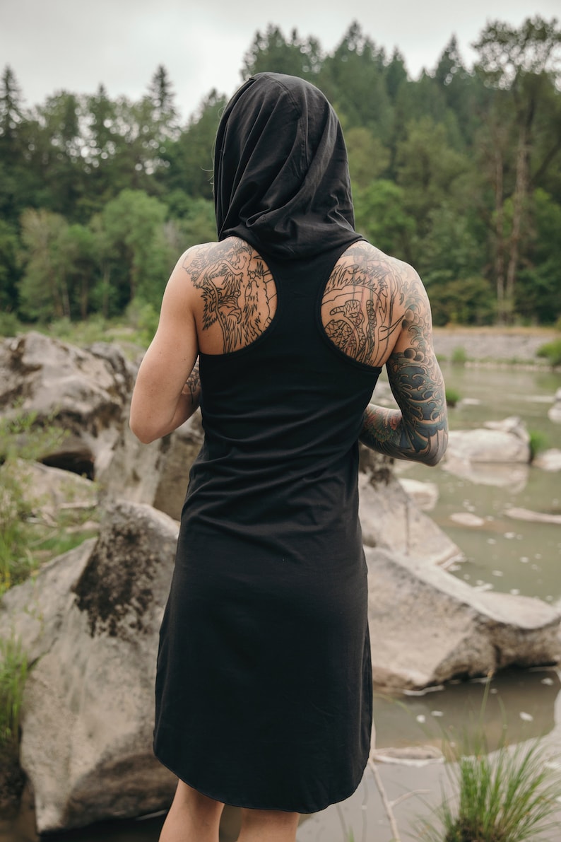 May include: A black hooded tank dress with a scoop back. The dress is made of a soft, comfortable fabric and has a relaxed fit. The model is wearing the dress with a hood pulled up over their head. The model has tattoos on their arms and back.