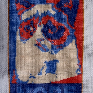 May include: Cross-stitch artwork featuring a cat's face in a pop art style. The design uses blue, red, and cream colors. The word "NOPE" is stitched in blue at the bottom of the artwork.