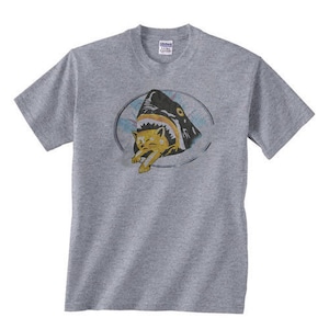 May include: A gray t-shirt with a graphic of a shark with a cat in its mouth. The graphic is in a vintage style with a yellow and black color scheme.