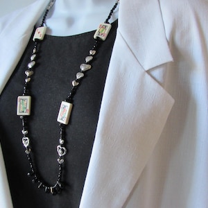 May include: A long necklace with black beads, silver heart-shaped charms, and rectangular pendants with colorful designs. The necklace is worn over a dark top and a white blazer.