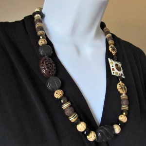 May include: A multi-colored beaded necklace with a square pendant featuring a geometric design. The necklace is made of brown, black, white, and gold beads.