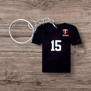 Könnte beinhalten: Schwarzer Trikot-förmiger Schlüsselanhänger mit der Nummer 15 in Weiß. Der Schlüsselanhänger hat eine silberne Kette und einen Ring. Das Trikot hat ein rot-weißes Logo und das Wort "TOLLGATE". Der Schlüsselanhänger liegt auf einer Holzoberfläche.