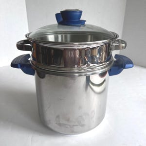 May include: A stainless steel multi-tier steamer pot with a clear glass lid. The pot features blue plastic handles on the sides and a matching blue knob on the lid. The design includes a deep base pot and a secondary steaming insert section.