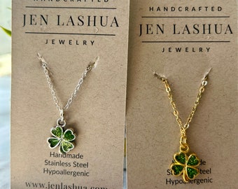 May include: Two necklaces with four-leaf clover charms on brown card stock. The necklaces are silver and gold. The card stock has the text "HANDCRAFTED JEN LASHUA JEWELRY" on it. The text "Handmade Stainless Steel Hypoallergenic www.jenlashua.com" is also on the card stock.