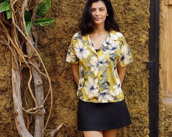 May include: A woman wearing a yellow floral print short-sleeved shirt, a black mini skirt, and black sandals. She is standing in front of a wall covered in moss.
