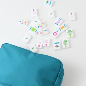 May include: A teal zippered pouch accompanied by a collection of white rectangular Mahjong tiles. The tiles feature colorful printed designs including numbers, bamboo stalks, wind directions, and joker icons in pink, green, blue, and orange.