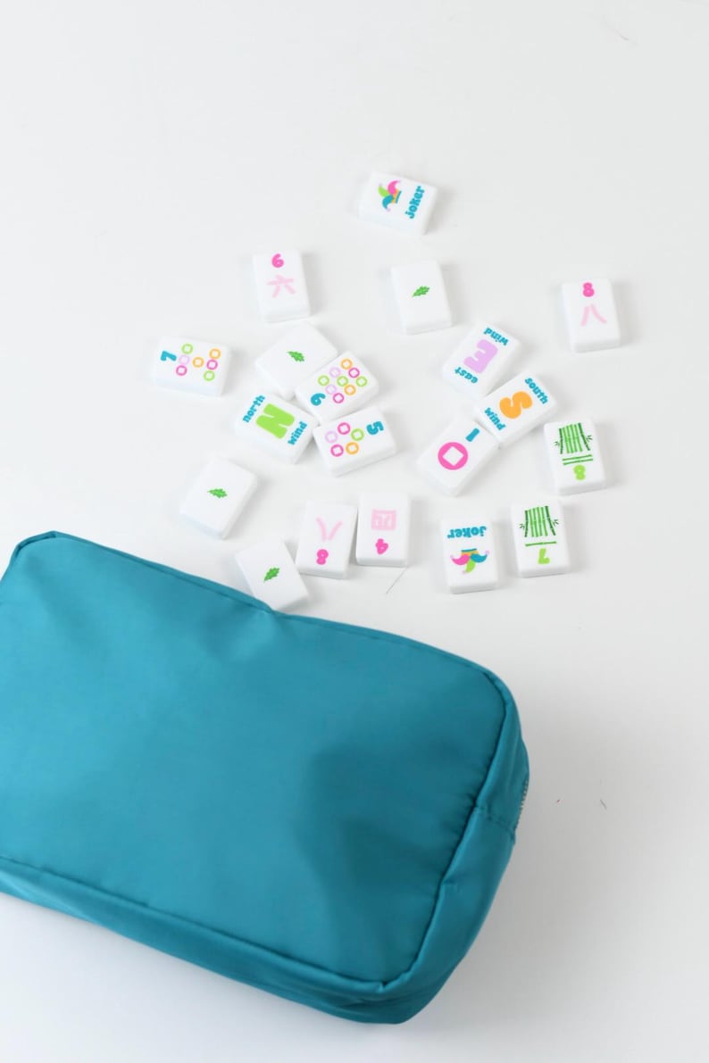 May include: A teal zippered pouch accompanied by a collection of white rectangular Mahjong tiles. The tiles feature colorful printed designs including numbers, bamboo stalks, wind directions, and joker icons in pink, green, blue, and orange.