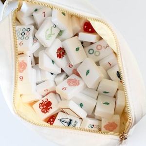 May include: A cream-colored zippered pouch filled with a collection of white rectangular mahjong tiles. The tiles feature various colorful printed designs including an octopus, a sailboat, seashells, leaves, numbers, and geometric patterns.