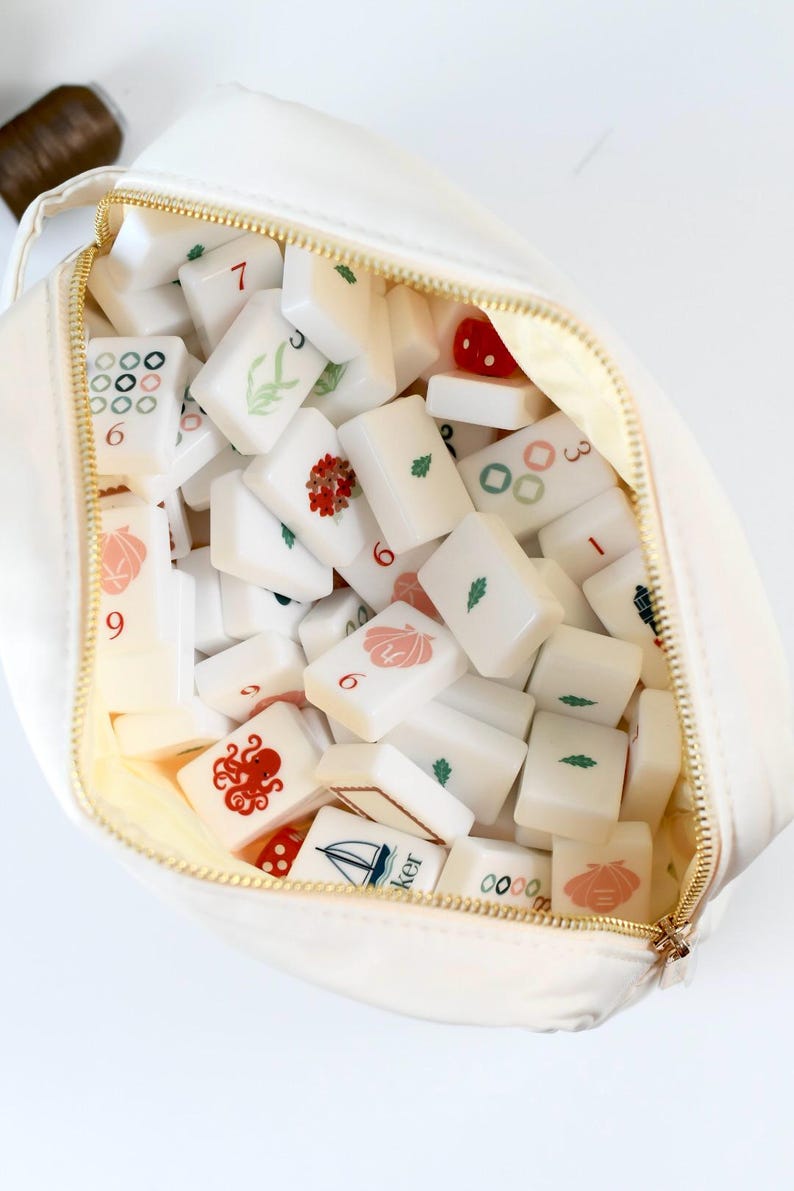 May include: A cream-colored zippered pouch filled with a collection of white rectangular mahjong tiles. The tiles feature various colorful printed designs including an octopus, a sailboat, seashells, leaves, numbers, and geometric patterns.