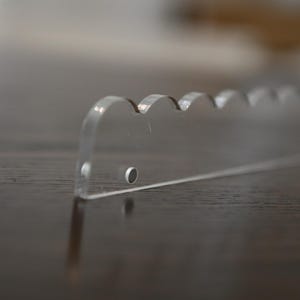 May include: A clear acrylic cake server with a scalloped edge. The server has a flat, rectangular shape with a series of rounded scallops along one side. The server is designed for cutting and serving cake.